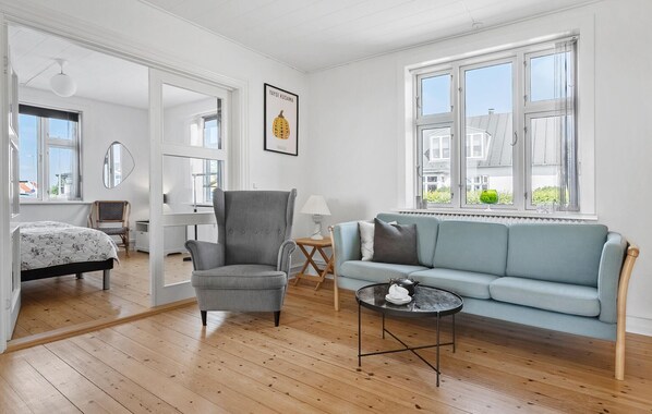 A room with a light blue sofa, a grey armchair, a small round table with a teapot, and a bed with a floral bedspread.