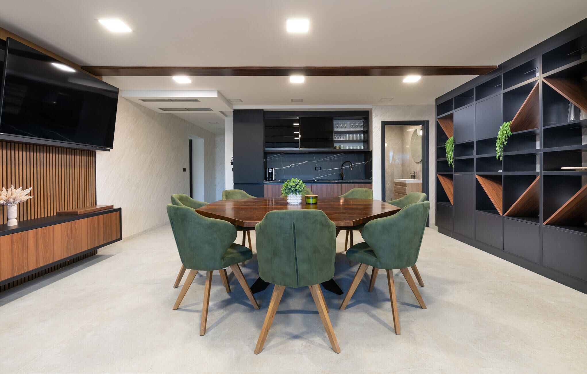 Un espace repas moderne avec une table en bois et des chaises à revêtement vert, une cuisine en arrière-plan et une étagère sombre agrémentée de plantes.