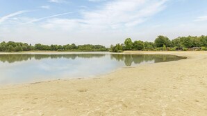 A calm lake with a sandy shore, surrounded by trees and a few buildings in the background. - Gorgeous home in Erm with house sea view (Erm)