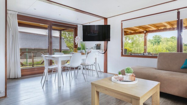 A modern living room with a wooden coffee table, a sofa, and a dining area with a white table and chairs. A flat-screen TV is mounted on the wall, and there are large windows offering a view of greenery. - Gorgeous home in Erm with house sea view (Erm)
