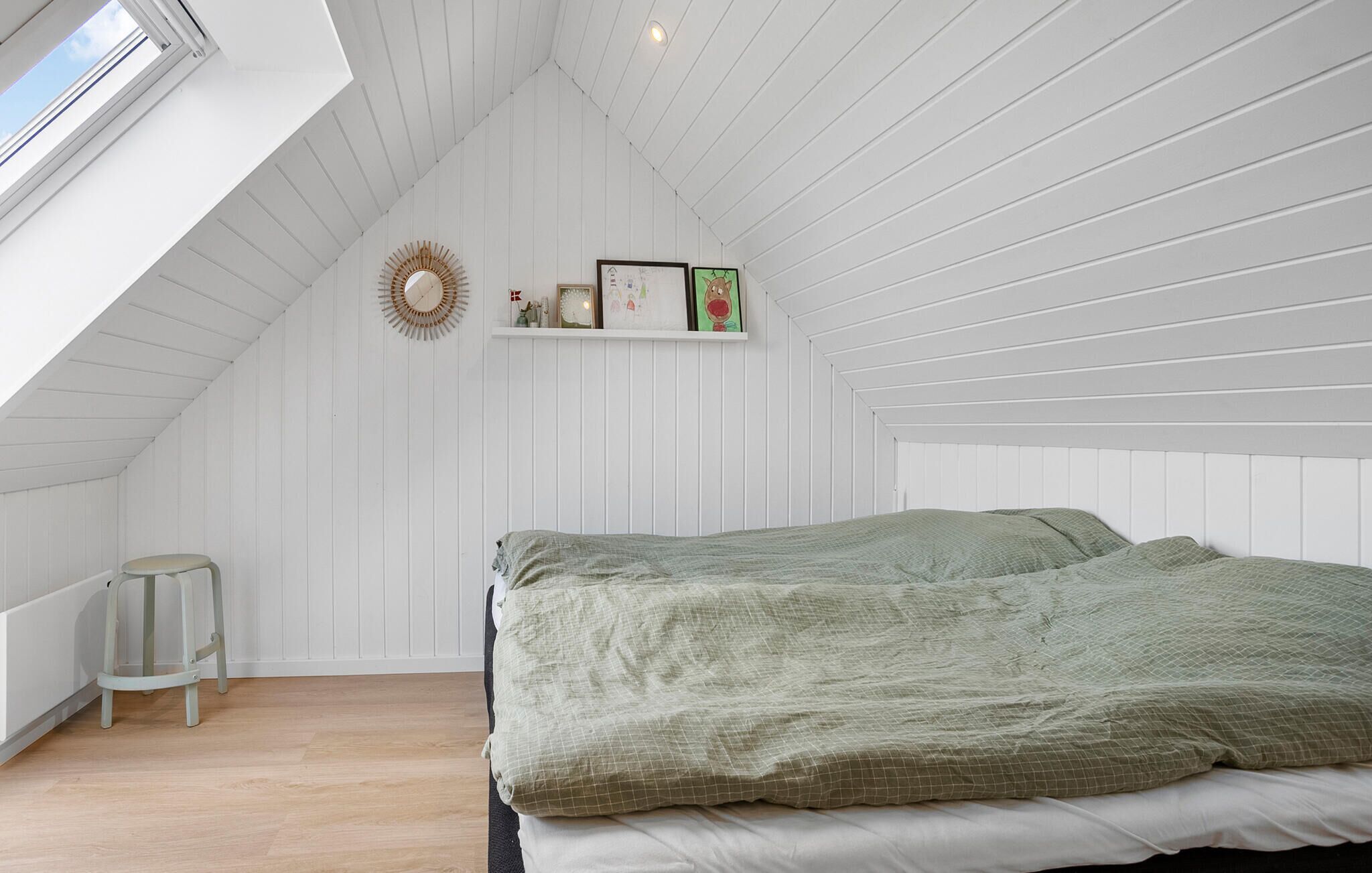 A bedroom with a sloped ceiling, a bed with green bedding, a small stool, and a shelf with framed pictures and a clock.