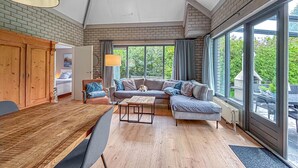 A cozy living room with a wooden dining table, a sofa, and a large window offering a view of greenery. - Nice home in Dirkshorn with WiFi (Dirkshorn)