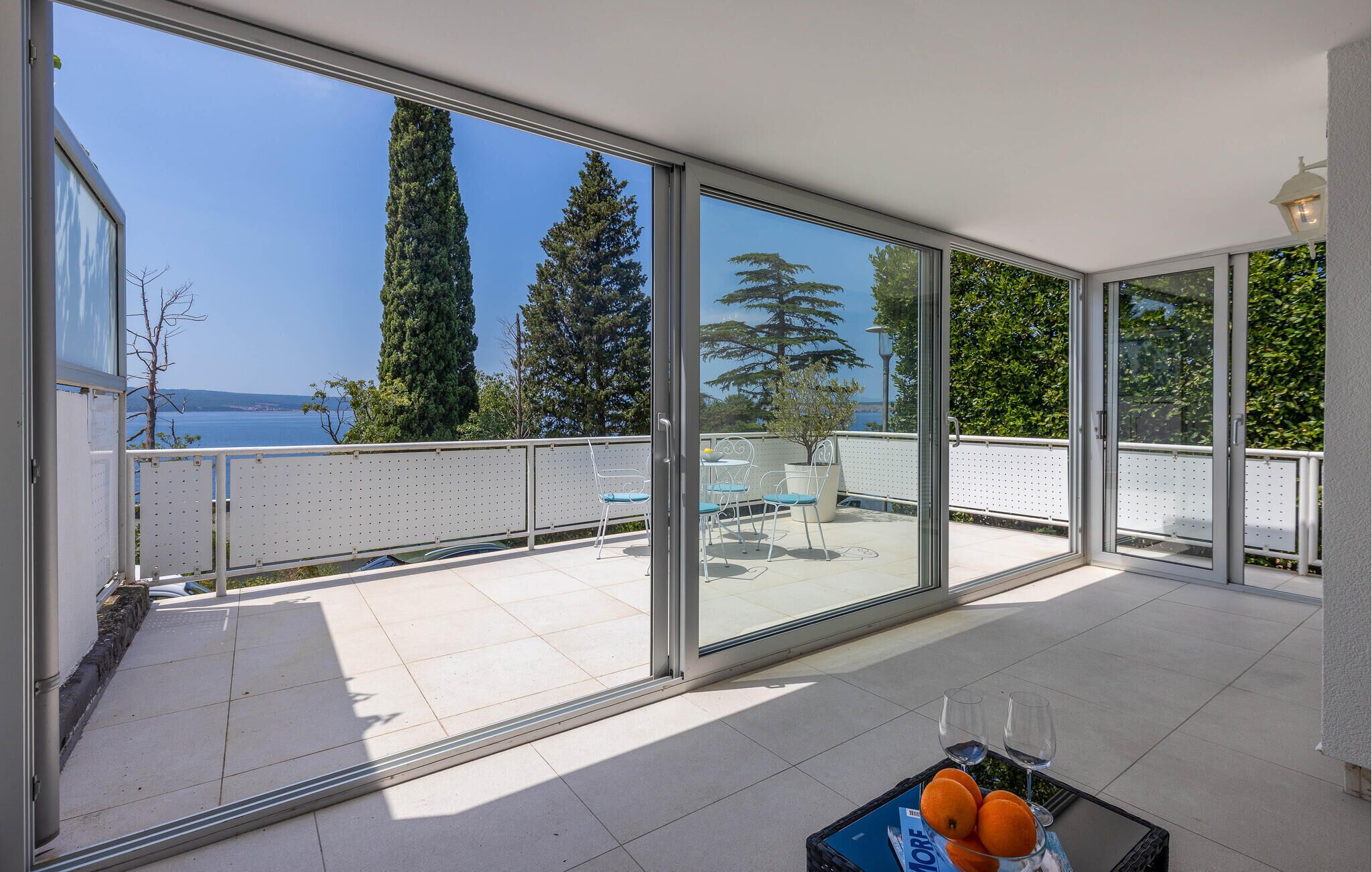 A spacious balcony with large sliding glass doors, offering a view of the sea and tall trees.