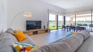 Een moderne woonkamer met een grote grijze bank, kleurrijke sierkussens, een flatscreen televisie en een eethoek met een tafel en stoelen.
