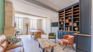 Een ruime woonkamer met een leren bank, een houten salontafel, een groene fauteuil en een oranje fauteuil. Er staat een boekenkast vol boeken en decoratieve objecten, en een open haard met een hoge, geweven vloerlamp.