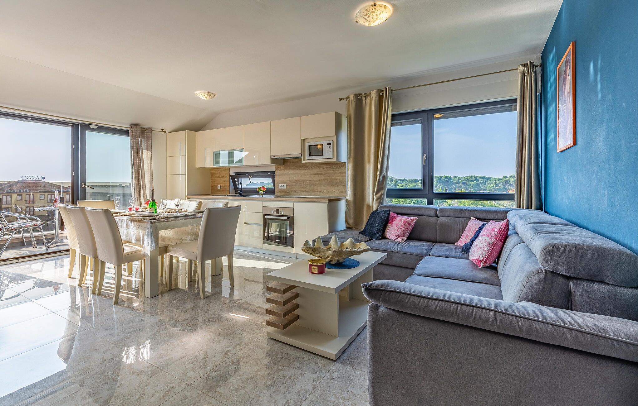 A modern living room with a grey sofa, a dining area with a table and chairs, a kitchen with white cabinets, and large windows offering a view of the city.