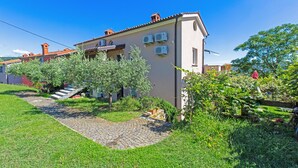 Ein zweistöckiges Haus mit gepflasterter Auffahrt, einem kleinen Garten mit Olivenbäumen und Klimaanlagen an der Außenwand.