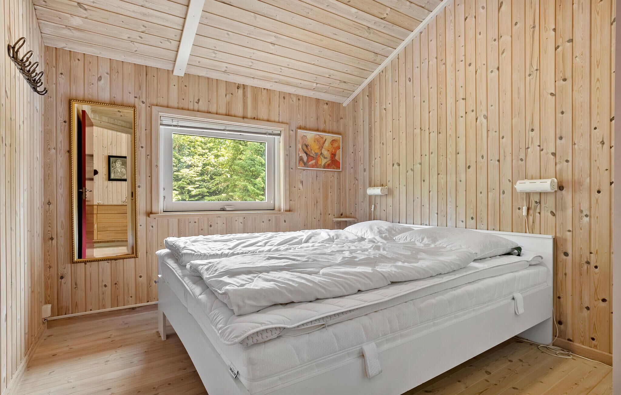 A bedroom with a wooden paneled wall, a window with a forest view, a bed with white bedding, and a mirror with a red frame.