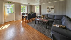 A spacious living area with a wooden floor, a dark grey sofa, a coffee table, and a dining table with chairs. There is a painting on the wall and a dog resting on the sofa. - Lovely apartment in Sperrebotn with WiFi (Sperrebotn)