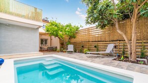 Ein rechteckiger Swimmingpool mit glasklar blauem Wasser, umgeben von einer gefliesten Terrasse. Zwei Liegestühle und ein Tisch mit Stuhl befinden sich im Schatten eines Baumes. Im Hintergrund sind eine Reetwand und ein Holzzaun mit Kletterpflanzen zu sehen.