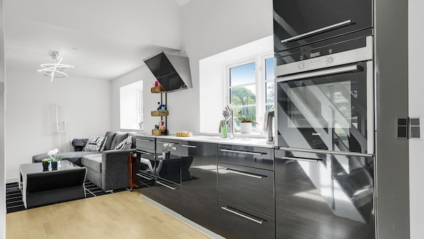 A modern kitchen with stainless steel appliances, a dining area with a grey sofa, and a window providing natural light.