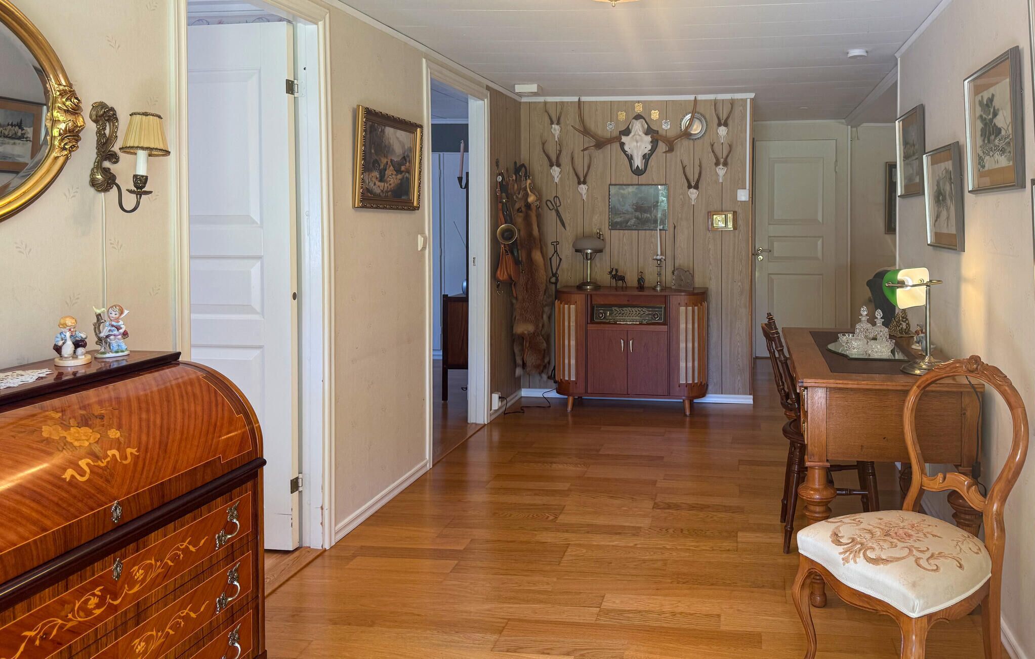 A room with wooden floors, a dining table with chairs, a cabinet, a mirror with a gold frame, and wall-mounted antler decorations.