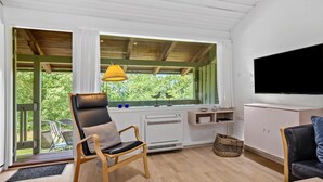 Een moderne kamer met een houten plafond, een groot raam met uitzicht op bomen, een zwarte leren fauteuil, een wit kastje en een televisie aan de muur.