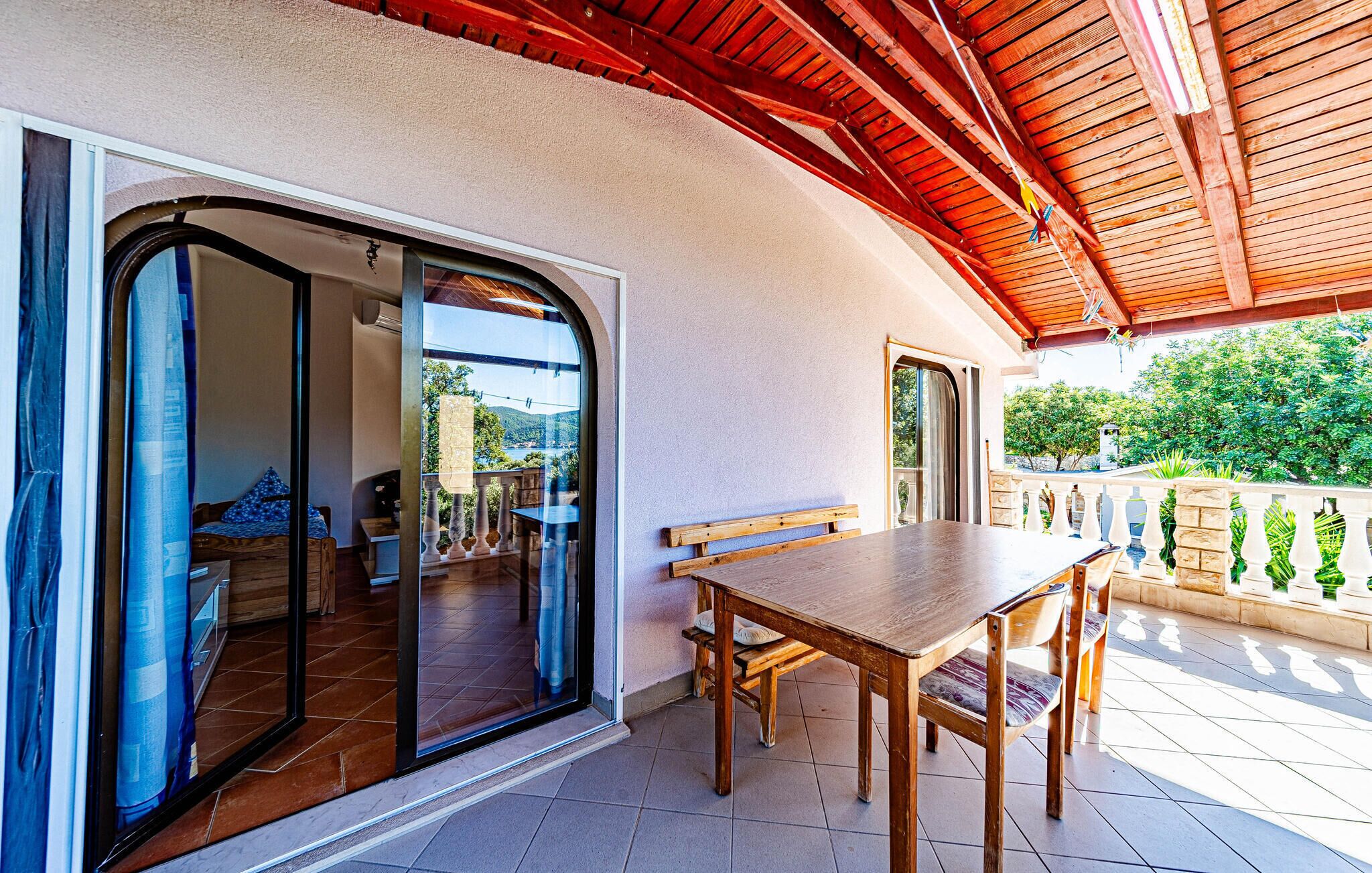 Un patio con una mesa y sillas de madera, un ventanal de vidrio que da a un espacio interior y vistas a zonas verdes y colinas.