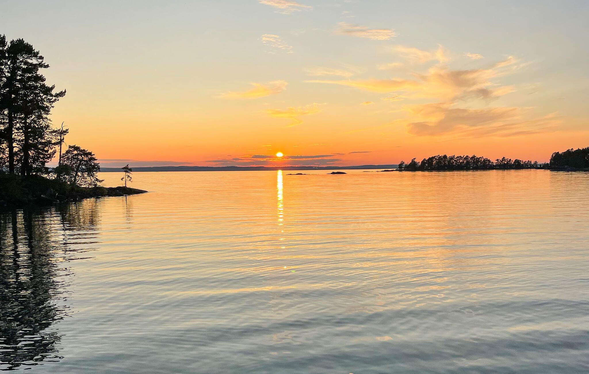 A calm lake at sunset with a clear sky, trees on the left, and a distant shoreline.