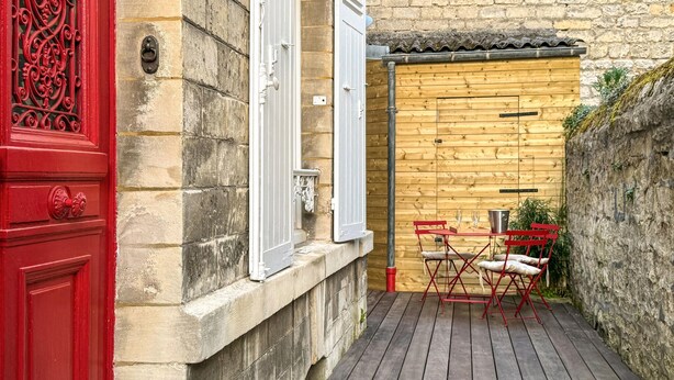 A wooden deck with a table and chairs, a red door, and a stone wall. - Charmante maison de ville (Chantilly)