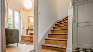 A staircase with wooden steps leading to a landing, a door with a white frame, and a view of a room with a large window and a hanging light fixture. - Charmante maison de ville (Chantilly)