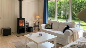 Un salon chaleureux avec un poêle à bois, un canapé blanc, une petite table et une vue sur un jardin verdoyant, visible à travers de grandes fenêtres.