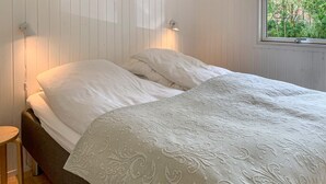Un lit avec du linge de lit blanc et une couverture à motifs, un tabouret en bois et une fenêtre donnant sur un paysage verdoyant.
