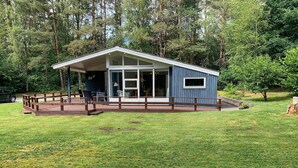 Une petite cabane à un seul étage, dotée d’un porche couvert et de grandes fenêtres, entourée d’une pelouse et d’arbres.