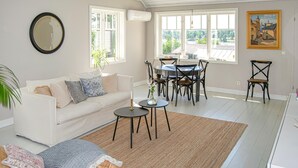 A living room with a white sofa, a dining table with chairs, a large window with a view, and a painting on the wall.