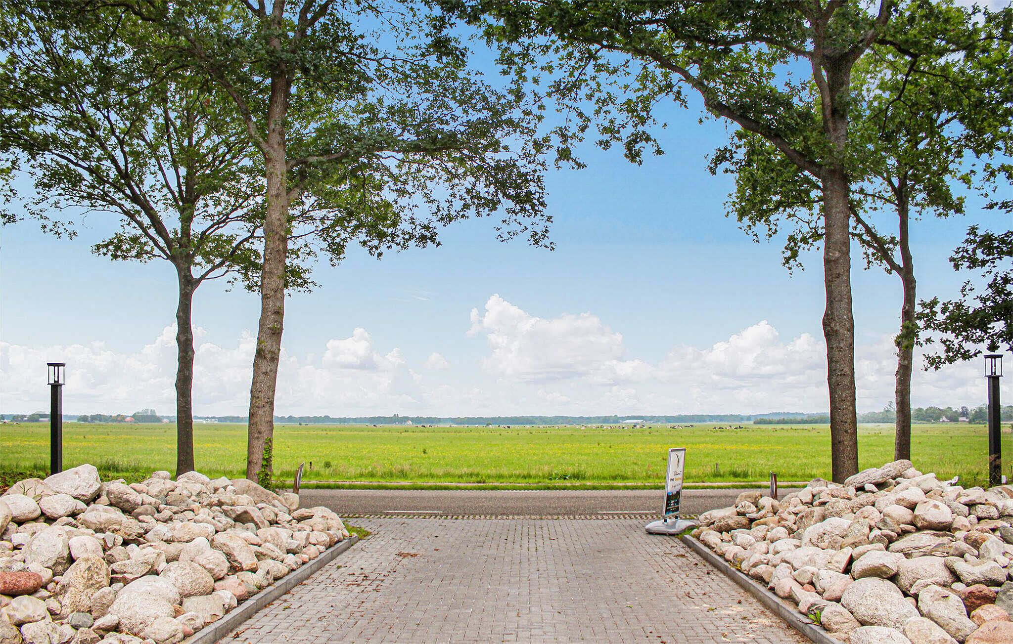 Un chemin pavé mène à un champ herbeux, bordé d’arbres de chaque côté, et on aperçoit un panneau indicateur au loin.