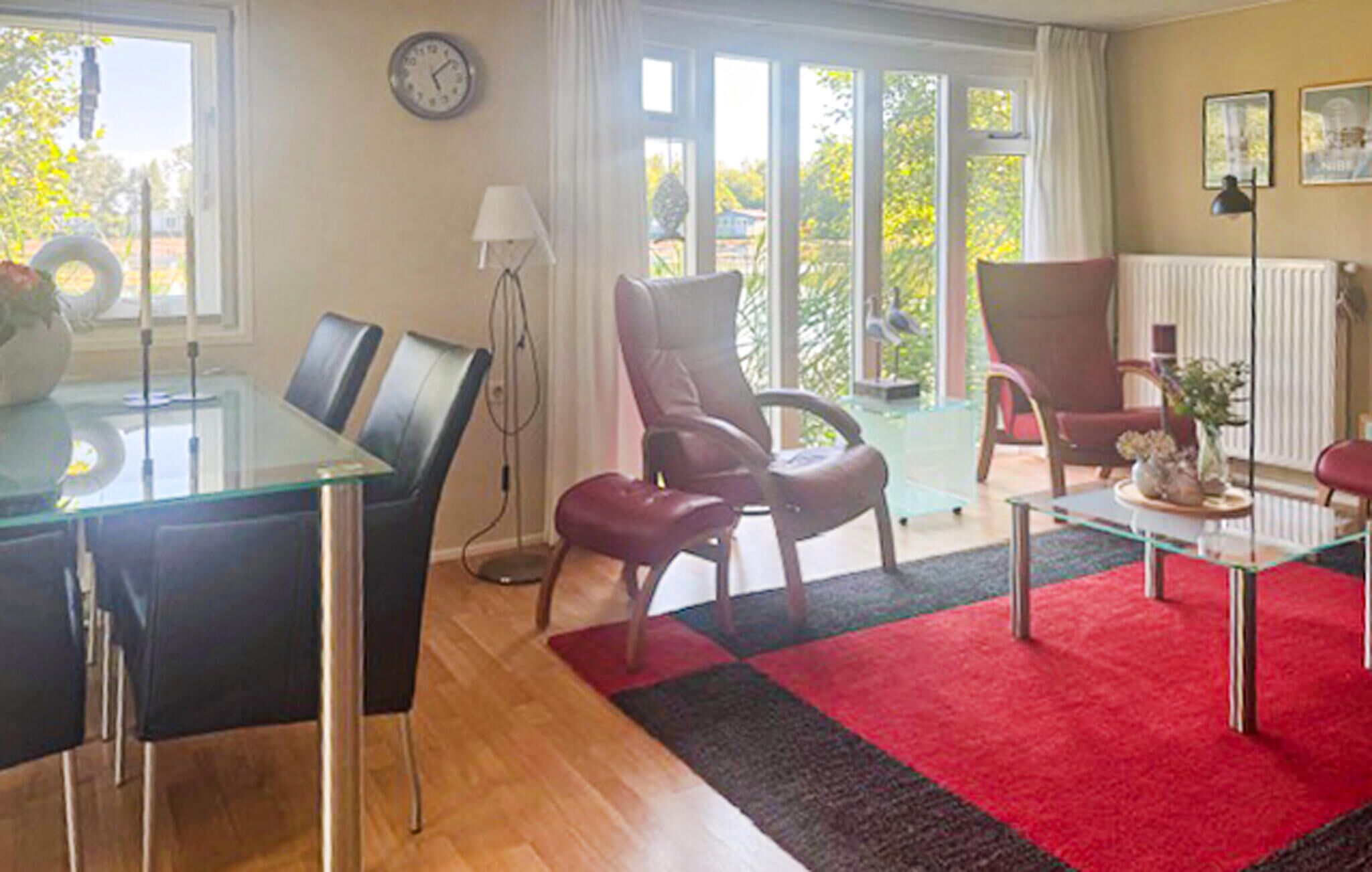 A modern living room with a glass dining table, two red chairs, a red and black rug, and a view of greenery through large windows.