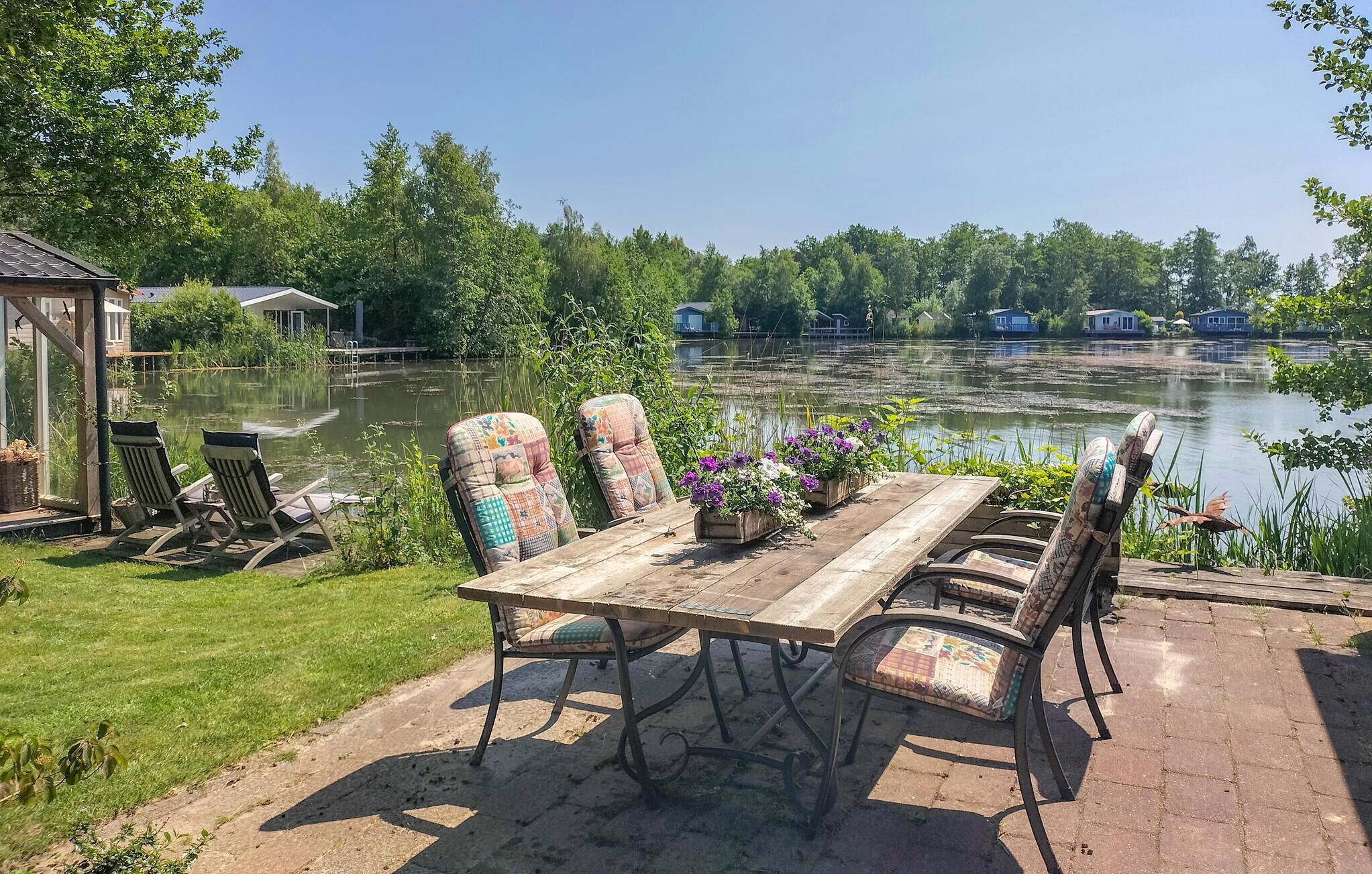 Une table en bois, entourée de chaises et d’un banc, se trouve au milieu d’un jardin d’où l’on aperçoit un lac et, au loin, des maisons.