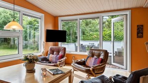 A cozy living room with two leather armchairs, a wooden coffee table, and a TV mounted on the wall. Large windows offer a view of the outdoors.