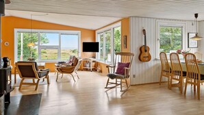 A cozy living room with a fireplace, comfortable seating, a dining area, and large windows offering a view of the outdoors.