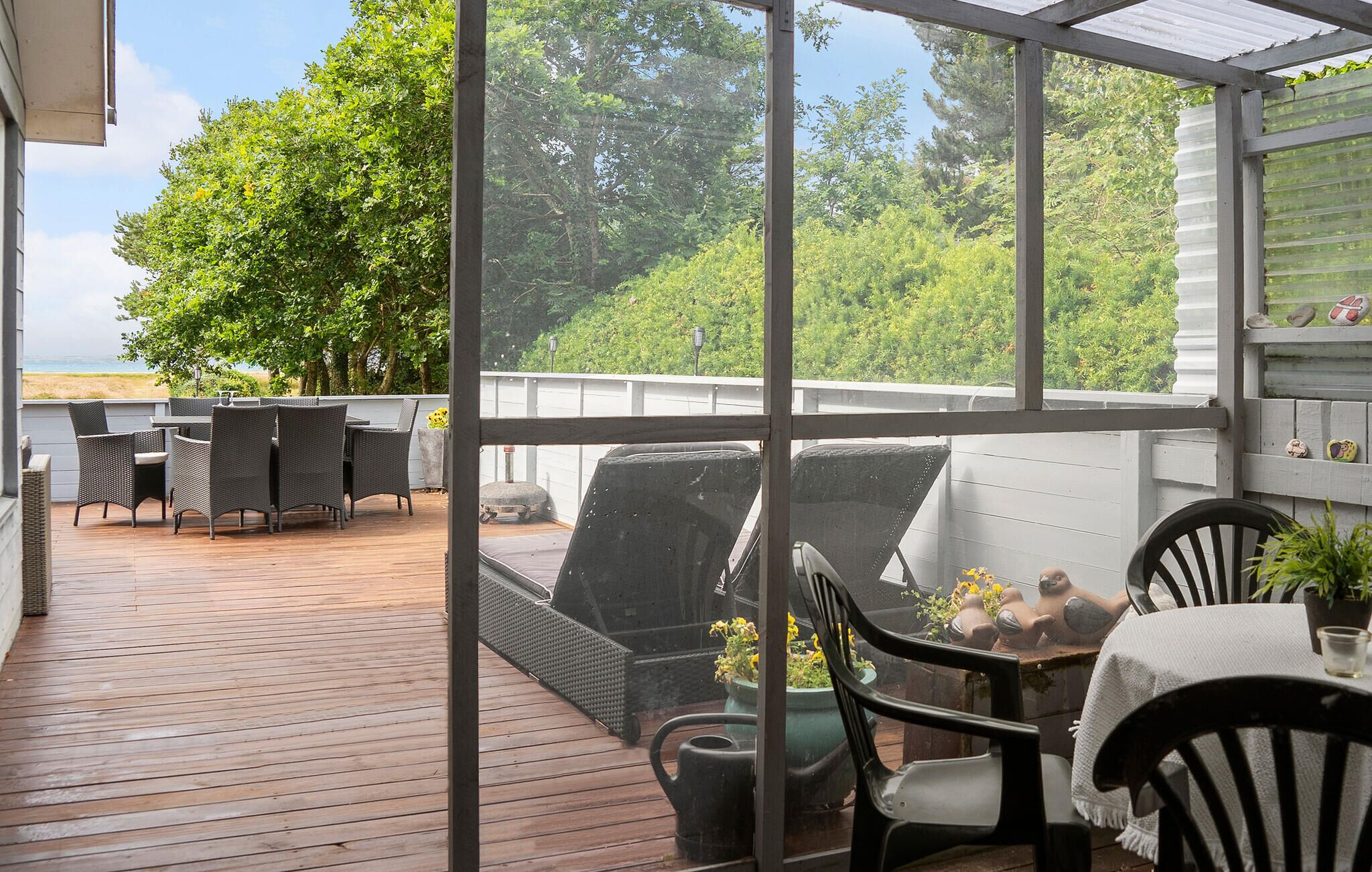 A covered patio with wicker furniture, a wooden deck, and a view of a beach and trees.