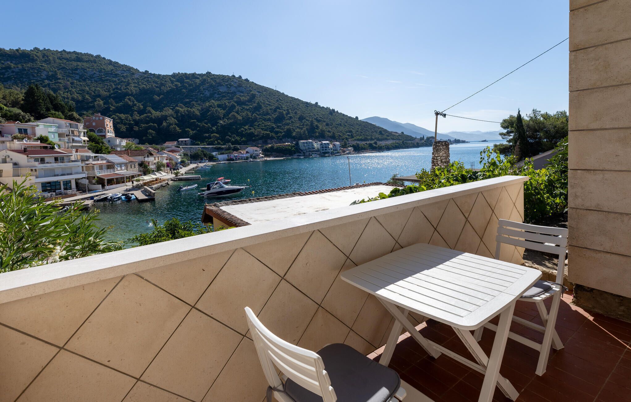 Un balcón con mesa y sillas que da a un puerto deportivo con barcos y un paisaje montañoso.