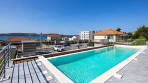 A swimming pool with a clear blue water, surrounded by a metal fence, and a view of the sea and buildings in the background. - Lovely apartment in Seget Donji with WiFi (Seget Donji)
