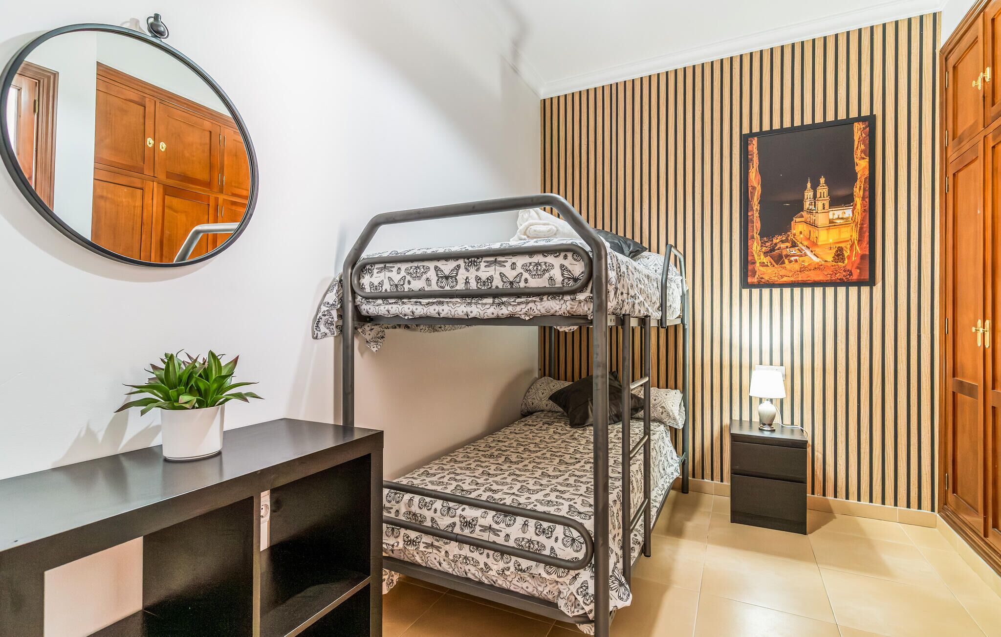 A bunk bed room with a wooden panel wall, a framed picture, a bedside table, and a potted plant.