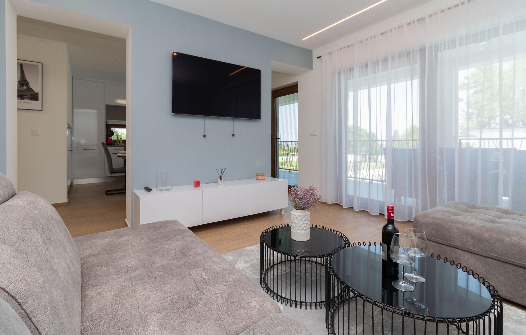 Ein modernes Wohnzimmer mit Fernseher, einem weißen Sofa, einem runden Glastisch und Blick auf einen Balkon durch transparente Vorhänge.