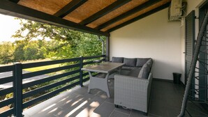 Eine überdachte Terrasse mit Tisch und zwei Korbssofas, einer Holzbalkendecke und Blick ins Grüne.