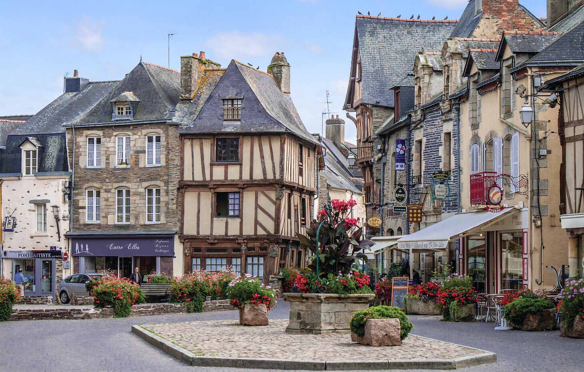 Une place pavée, bordée de bâtiments historiques, avec un parterre central et des espaces aménagés pour s’asseoir à l’extérieur.