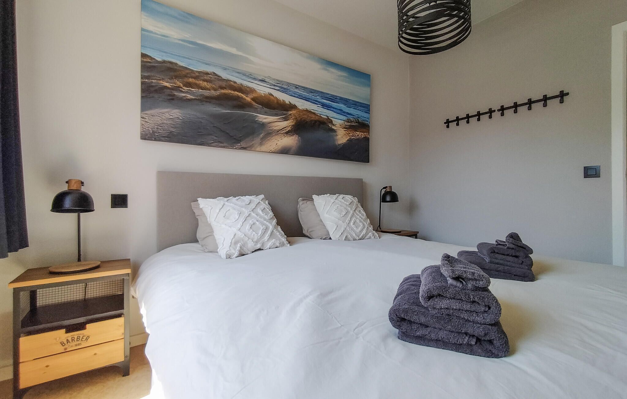 Ein Schlafzimmer mit einem großen Bett, einem Nachttisch, einer Lampe, einem Strandbild und einer Wandleuchte.