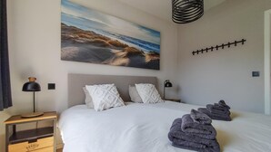 A bedroom with a large bed, a nightstand, a lamp, a painting of a beach, and a wall-mounted light fixture. - Pet friendly apartment in Oostende with WiFi (Oostende)