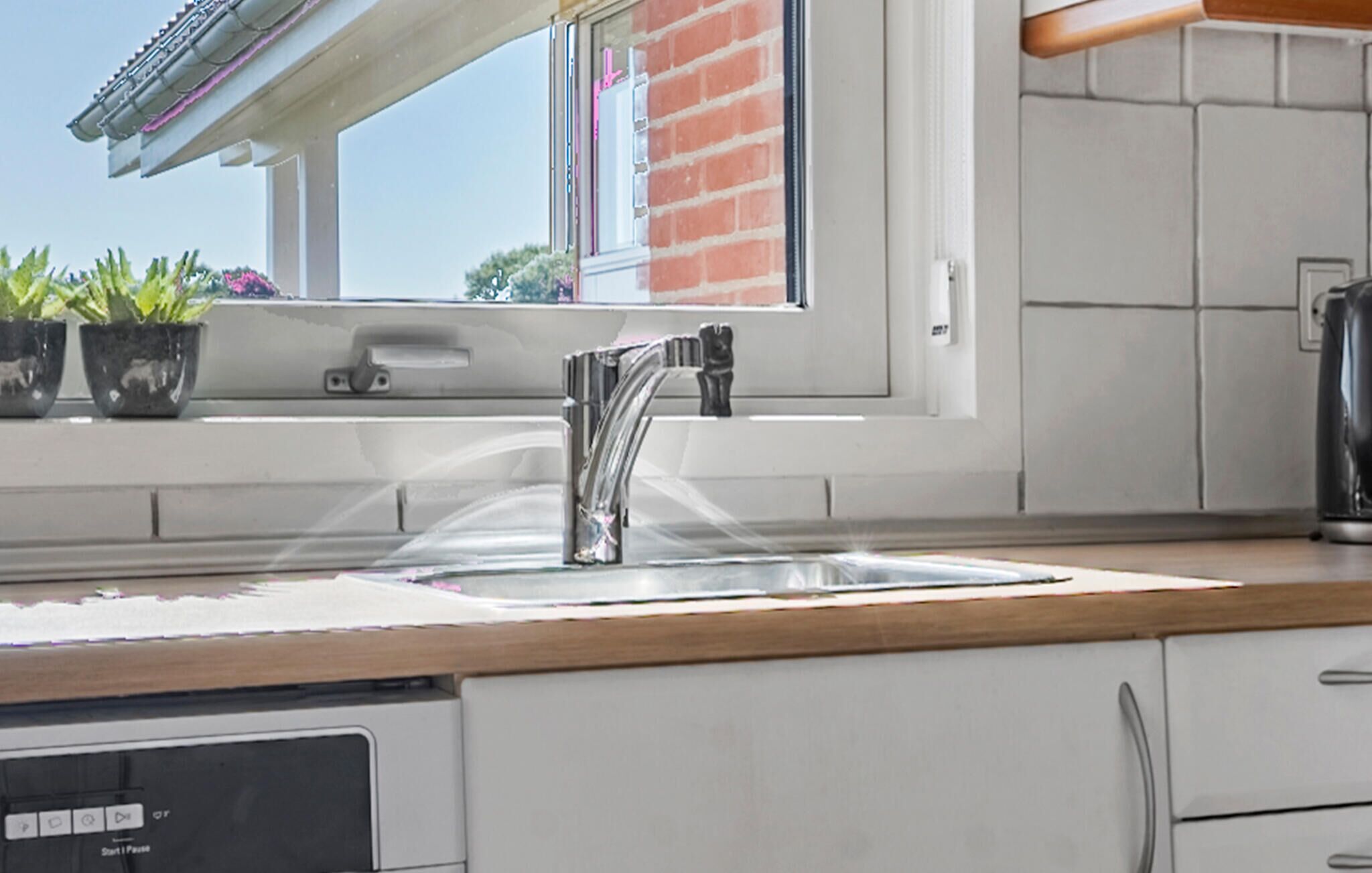 A kitchen with a stainless steel faucet, a window with a view of a brick building, and a dish drying rack.