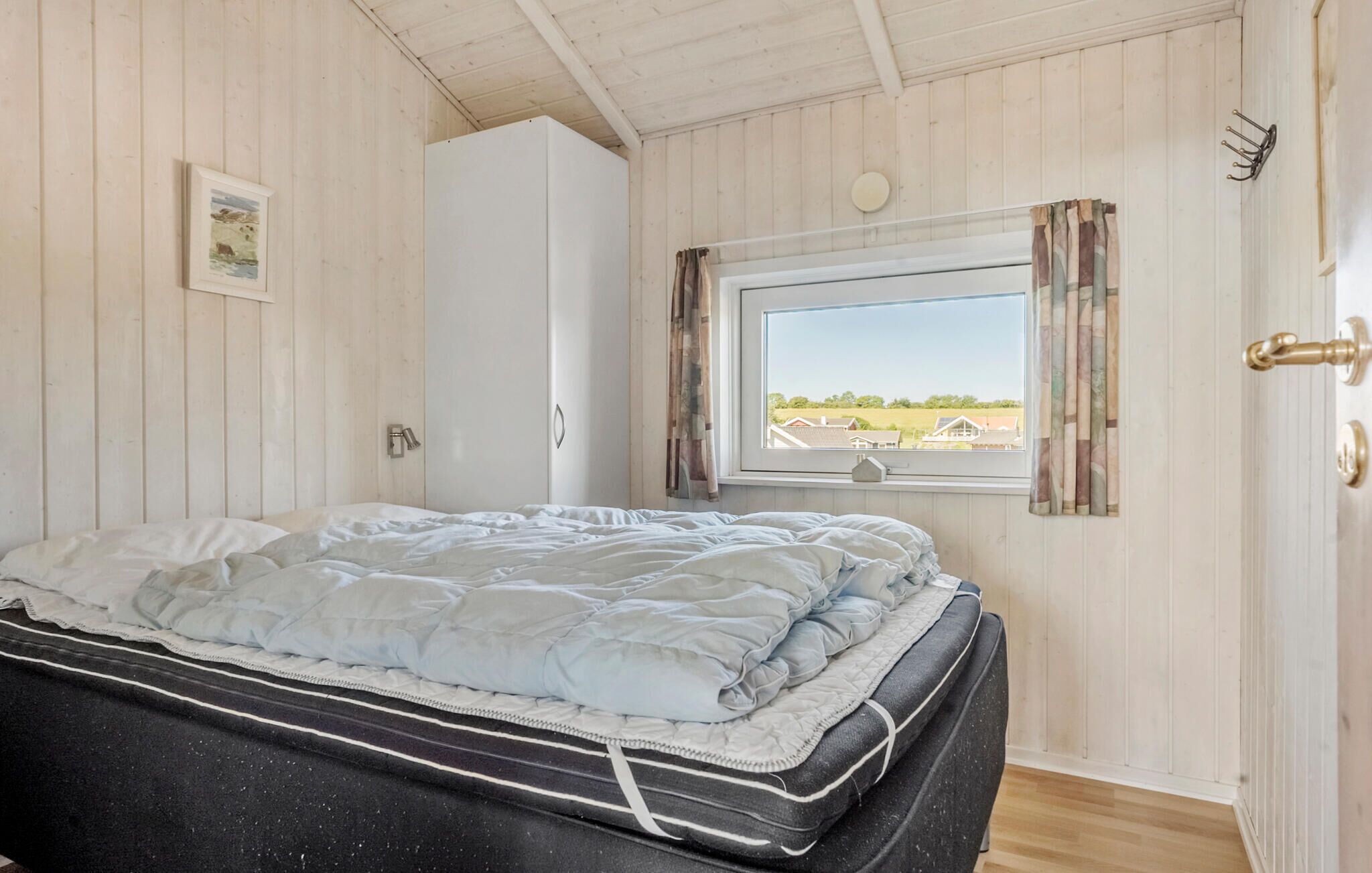 A bedroom with a bed, a window with curtains, a door with a handle, and a wall-mounted light fixture.