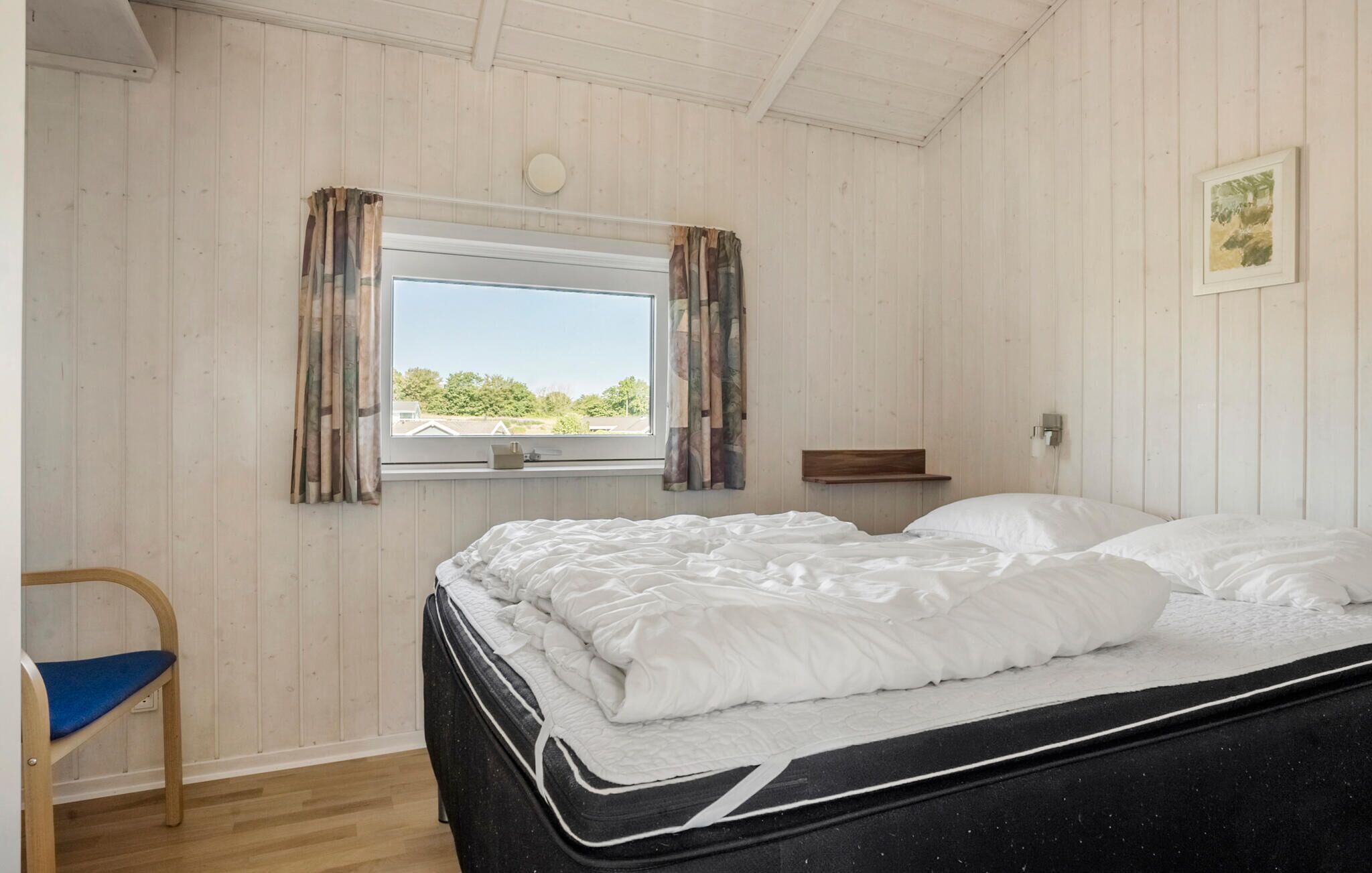 A bedroom with a bed, a chair, a window with curtains, a bedside table, and a picture on the wall.