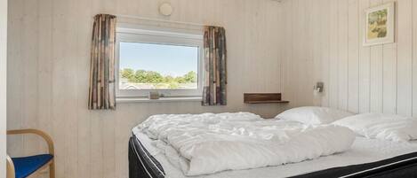 A bedroom with a bed, a chair, a window with curtains, a bedside table, and a picture on the wall.