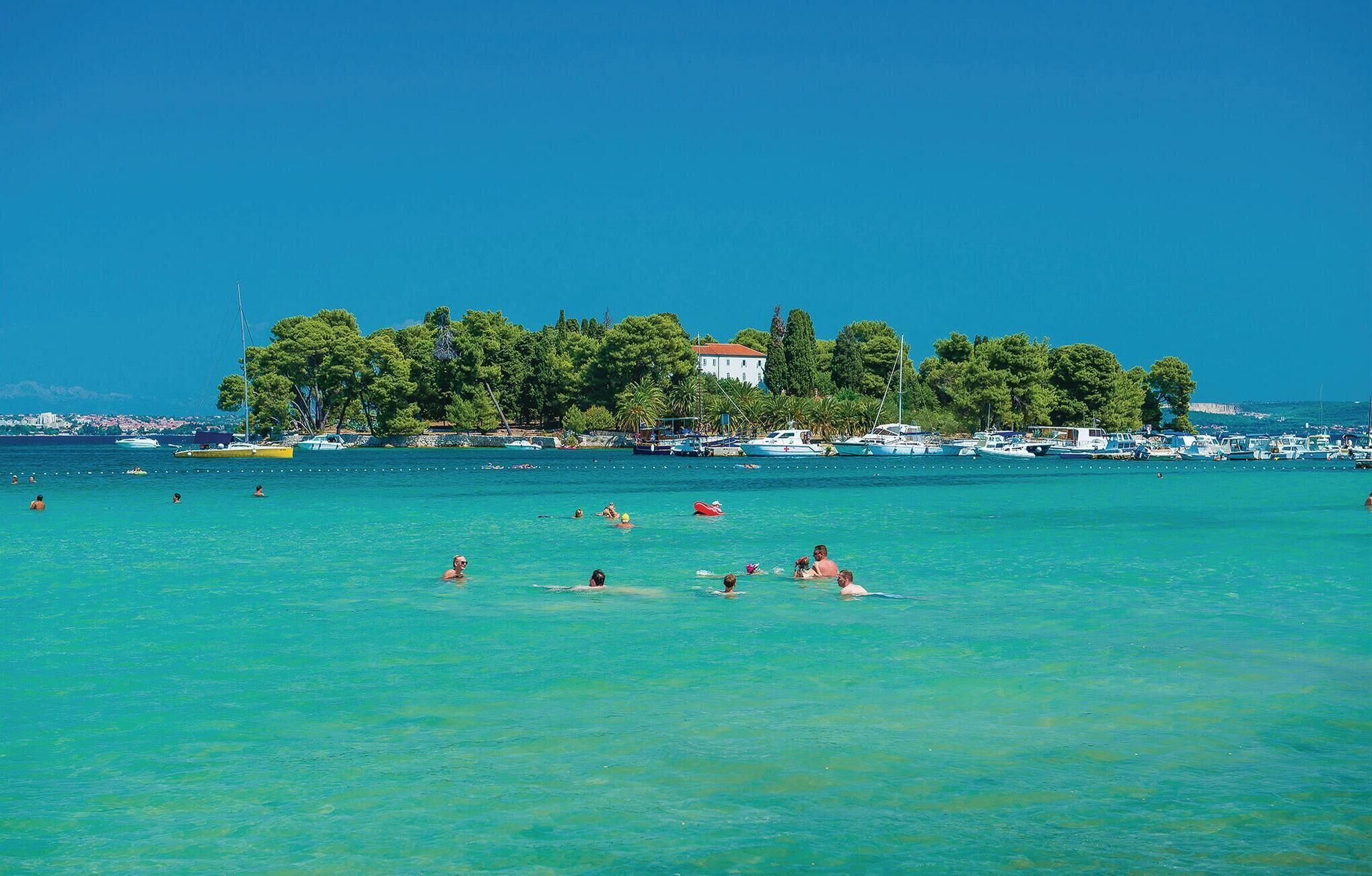 A coastal scene with clear turquoise waters, a row of boats, and a building with a red roof.