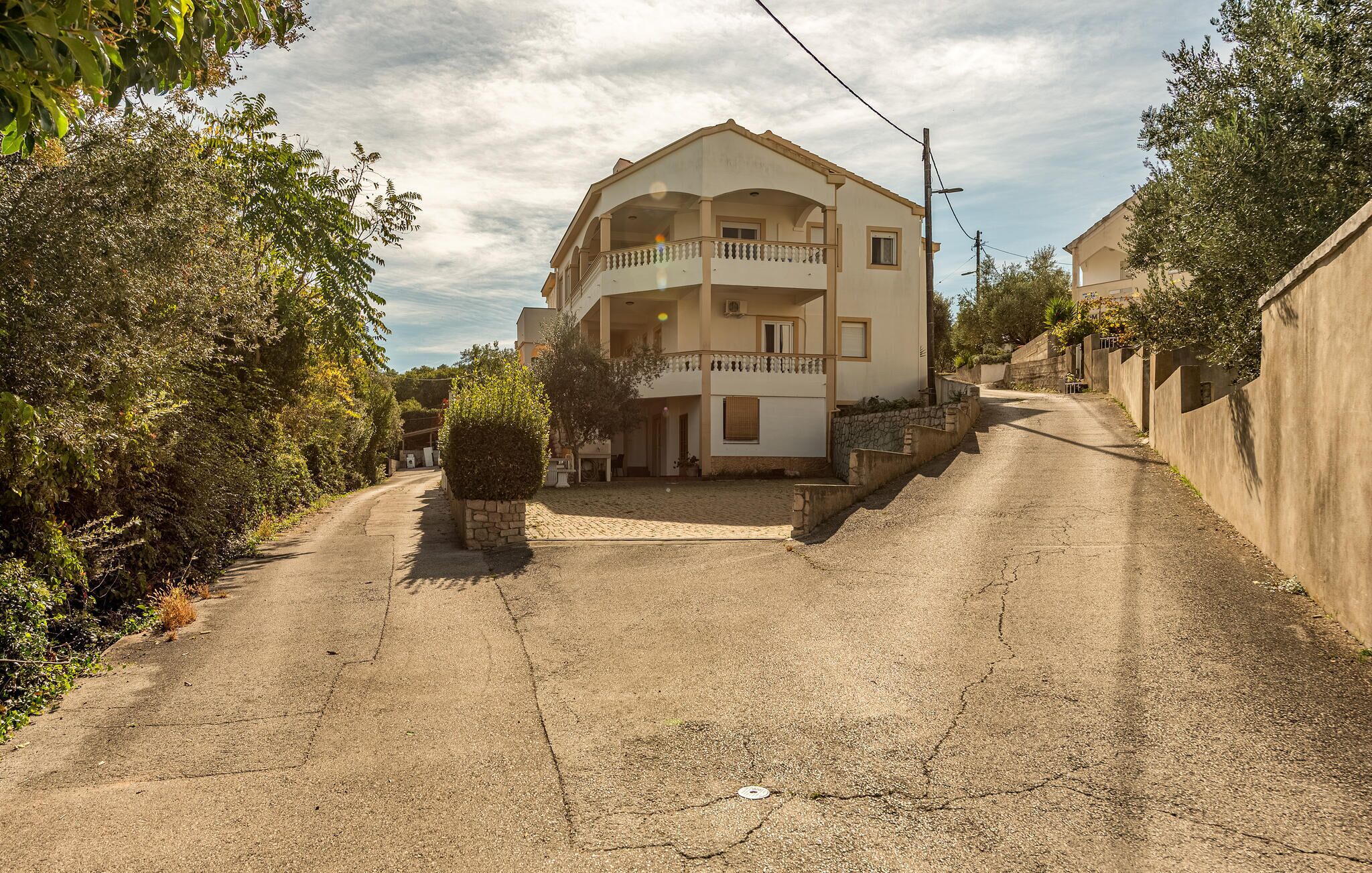 Un edificio a due piani con un balcone, circondato da alberi e un vialetto sterrato.