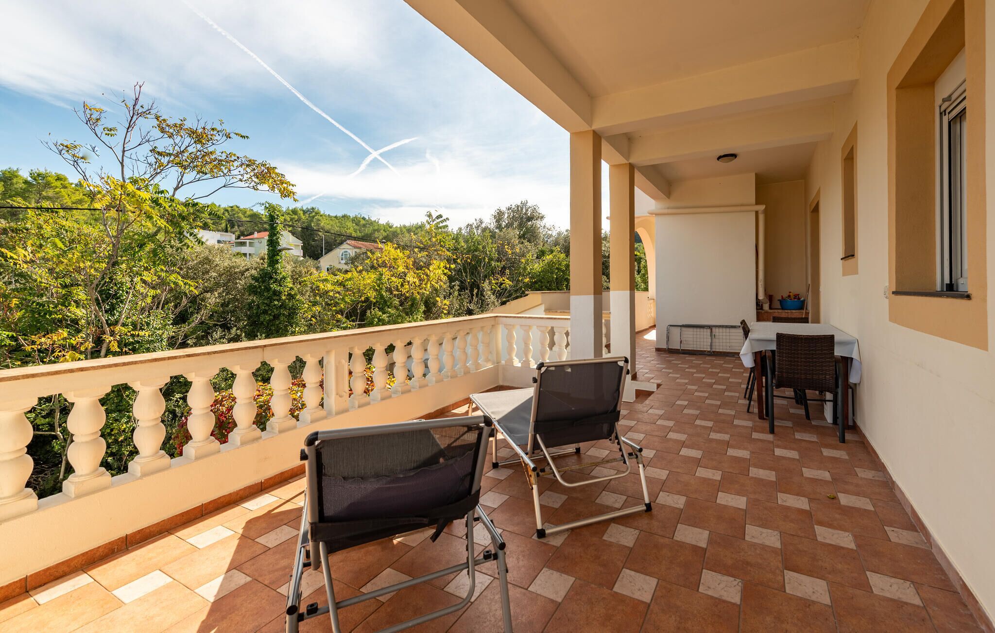 A balcony with two chairs, a table, and a view of greenery and houses.