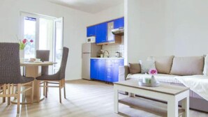 Een woonkamer met een bank, een salontafel en twee stoelen. Er is een keukenhoek met blauwe kasten en een raam.