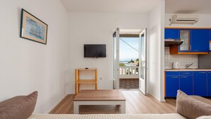 Een slaapkamer met een bed, nachtkastje en televisie. Er is een balkon met uitzicht op zee en een keuken met blauwe kastjes.