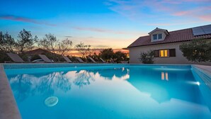 Une piscine à l’eau d’un bleu cristallin, entourée de chaises longues et bordée d’un bâtiment dont le toit est recouvert de tuiles.