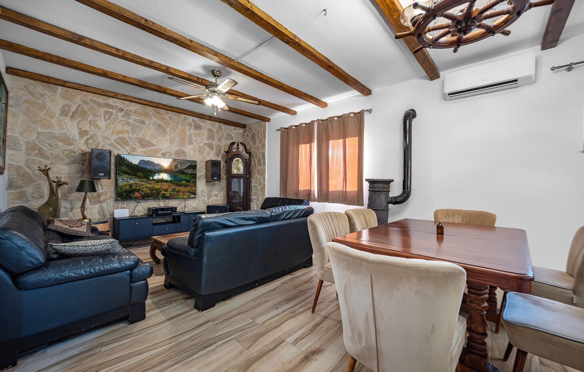 A living room with a stone wall, wooden beams, a fireplace, a dining table with chairs, and a television.
